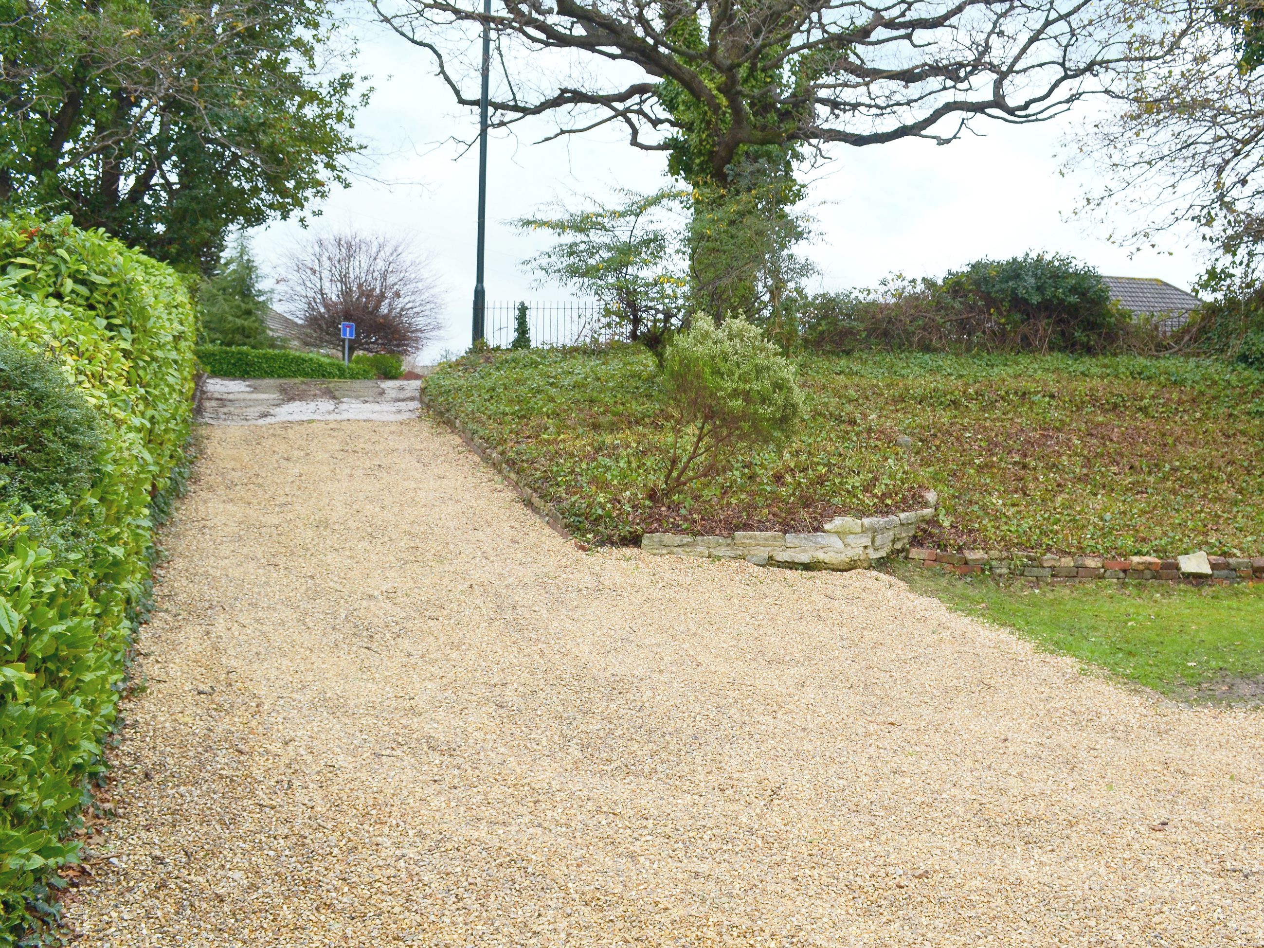 A gravel path flanked by green hedges and a stone wall, leading to a building with a tiled roof.