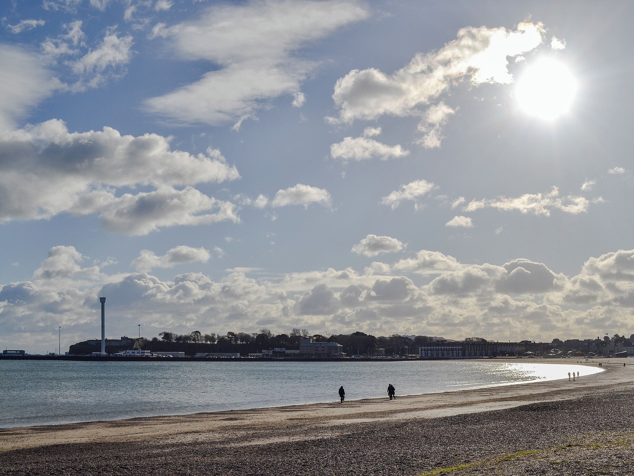 A beach with a lighthouse in the background, a clear sky, and a few people walking along the shore.