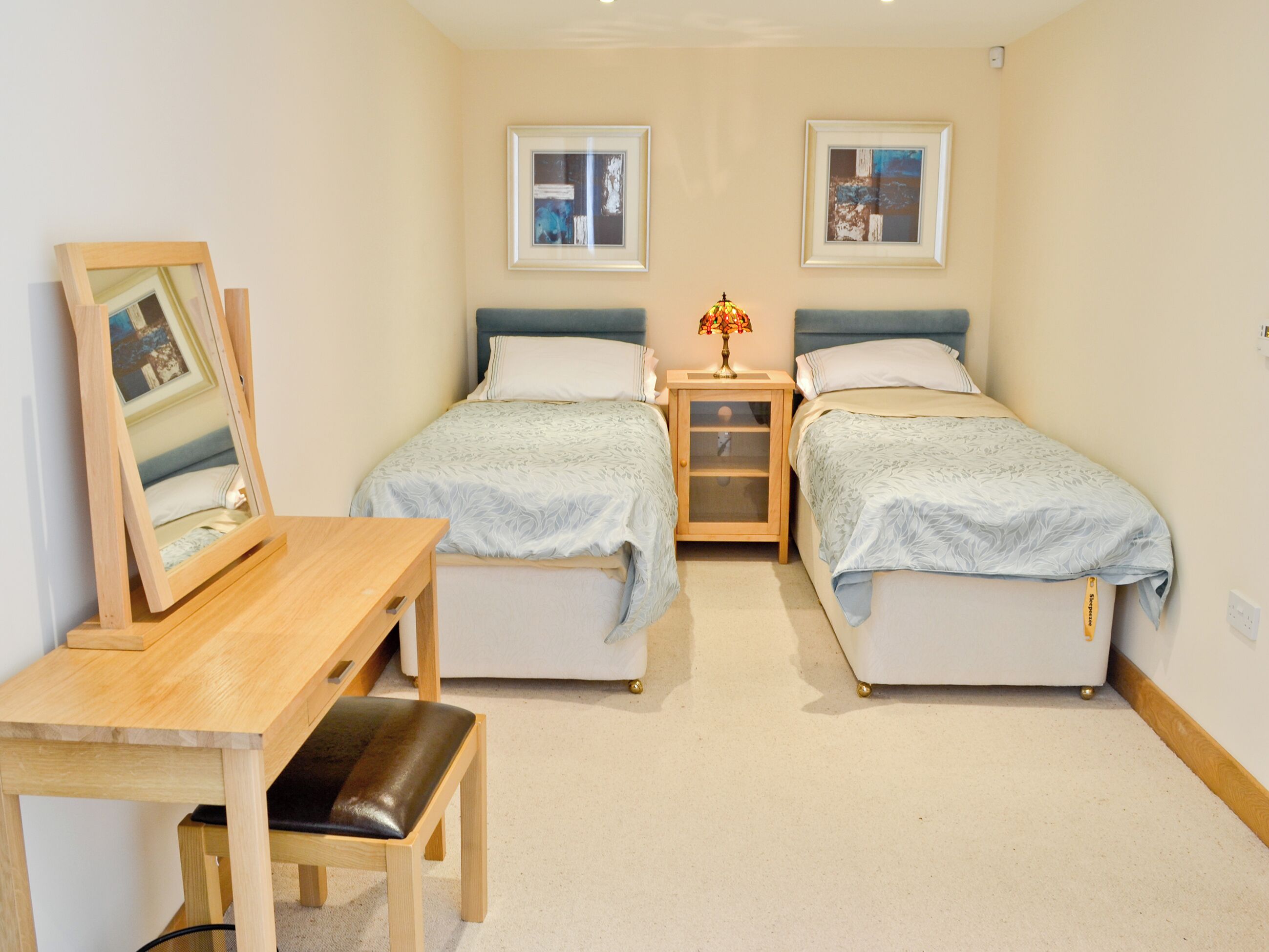 Un dormitorio con dos camas individuales, un escritorio de madera, una silla, una mesita de noche con una lámpara y dos cuadros en la pared.
