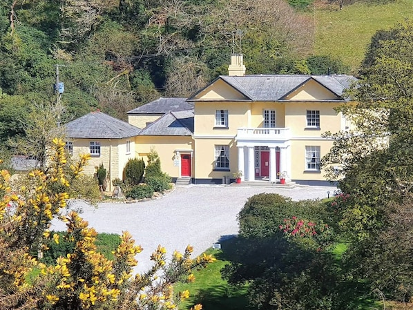 A large, yellow building with a red entrance, surrounded by greenery and a gravel driveway.