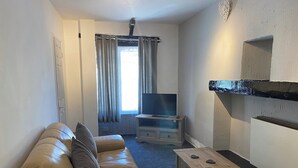 Un salon comprenant un canapé, une petite table, un téléviseur posé sur un meuble, une fenêtre avec des rideaux et une étagère fixée au mur.