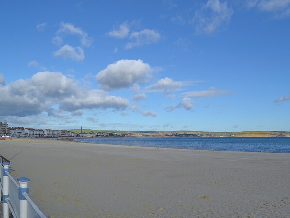 A coastal view with a sandy beach, a clear blue sky, and a distant town with buildings and hills.