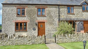 A stone cottage with a wooden door, two windows with shutters, and a stone wall with a gate.