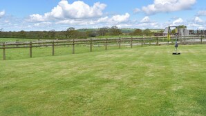A grassy field with a wooden fence and a tennis court with a net and ball machine.
