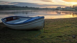Ein kleines Boot liegt an einem grasbewachsen Ufer eines stillen Sees, im Hintergrund ein Sonnenuntergang.