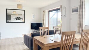 A modern living room with a wooden dining table, comfortable seating, a flat-screen TV, and a large window with curtains. - 2 bedroom accommodation in Padstow (Padstow)
