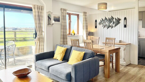 A modern living room with a grey sofa, wooden dining table, and chairs. There is a large window with curtains, a kitchen area, and a view of the countryside. - 2 bedroom accommodation in Padstow (Padstow)