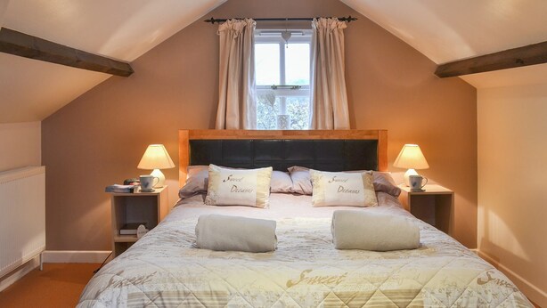 A bedroom with a large bed, two bedside tables, a window with curtains, and a wooden ceiling beam.