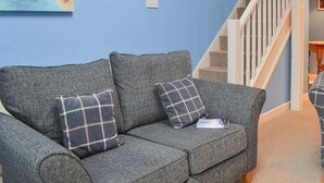 A grey patterned sofa with two plaid cushions, a painting of a coastal scene, and a staircase with white railings.