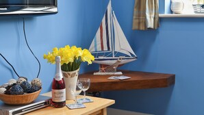 A room with a blue wall, a television mounted on the wall, a wooden table with a bottle of wine, two glasses, a basket of decorative eggs, and a small sailboat model.