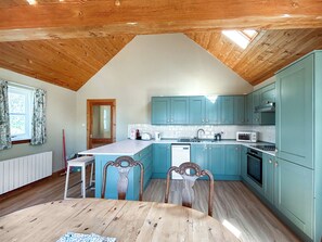 A kitchen with wooden ceiling beams, light blue cabinets, a dining table with chairs, and a window with curtains.
