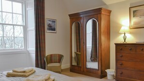 Une chambre à coucher avec un lit, une armoire en bois, une chaise, une lampe et une fenêtre avec des rideaux.