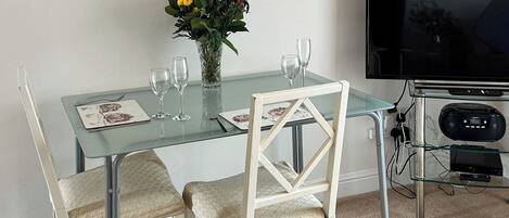 A dining area with a glass table, two chairs, a vase with flowers, and a television.