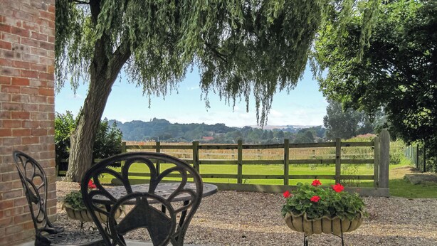 A patio with two wrought iron chairs and a table, a brick wall, and a view of a green field and trees in the background. - 1 bedroom accommodation in Little Tathwell, near Louth (Little Tathwell, near Louth)