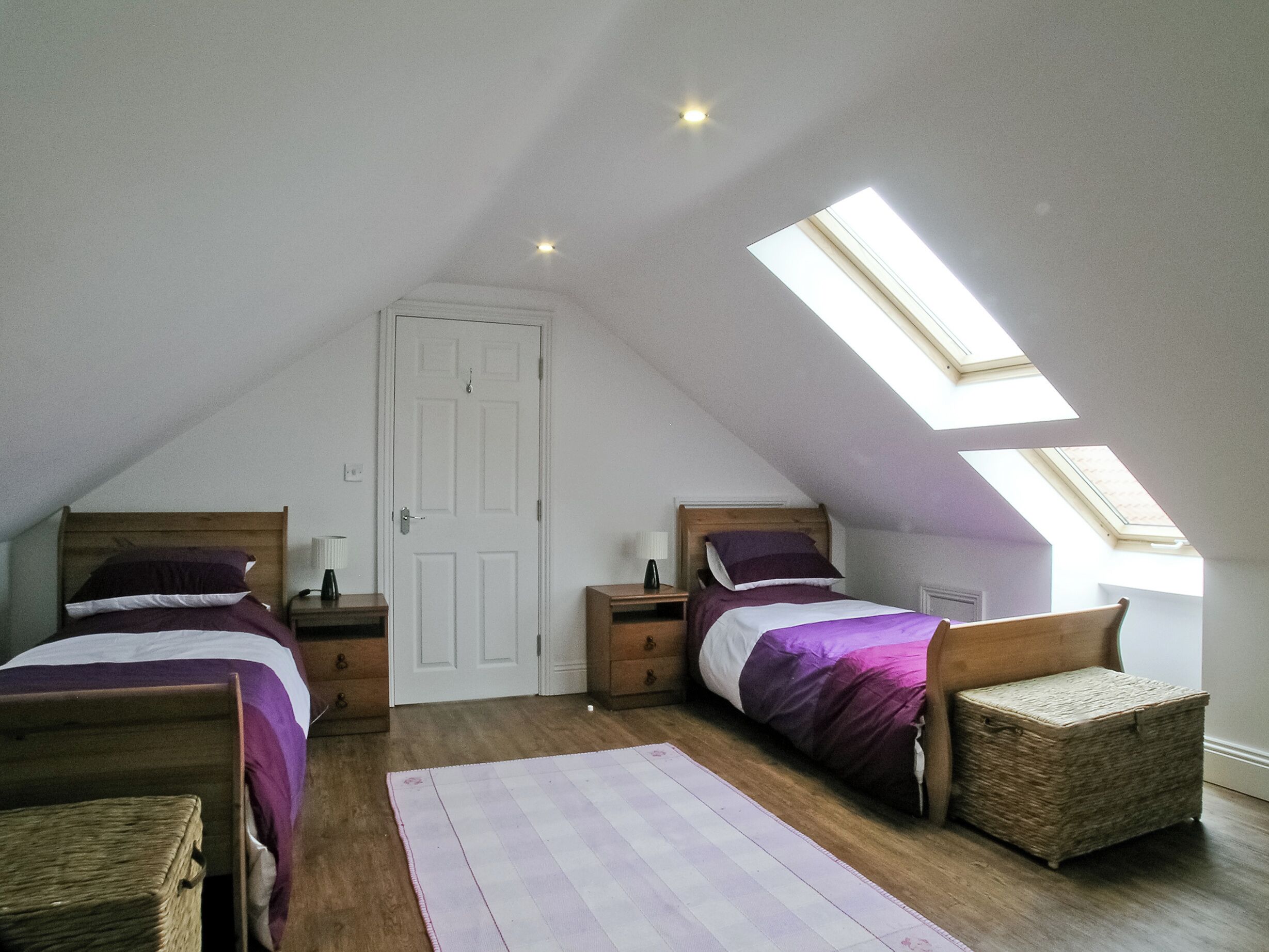 Une chambre avec deux lits, un puits de lumière, un plancher en bois et une porte blanche.