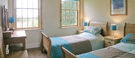 A bedroom with two beds, a wooden dresser, a desk, and two windows with blue curtains.