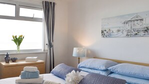Ein ordentlich bezogenes Bett mit blau-weißer Bettwäsche, ein Nachttisch mit Lampe, eine Vase mit Blumen und ein Strandmotiv als Bild an der Wand.