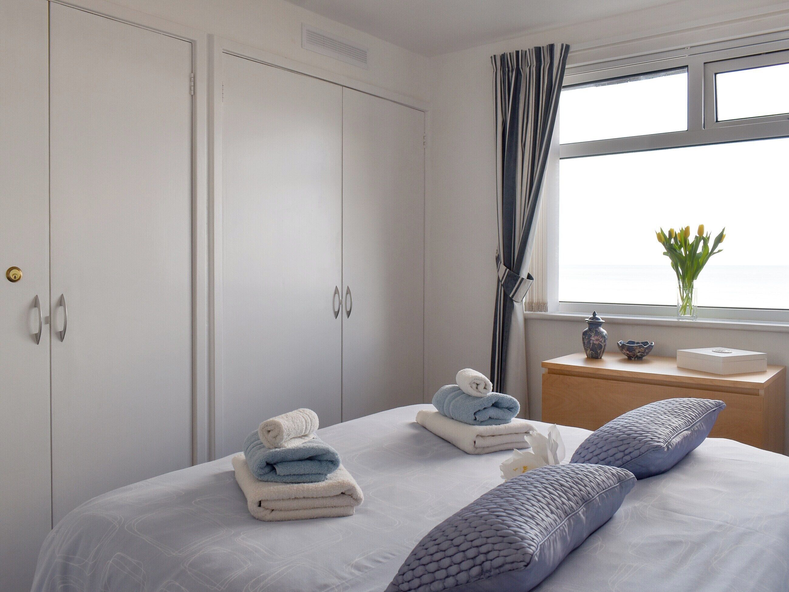 Un dormitorio con cama, mesita de noche, ventana con cortinas y vista al mar.
