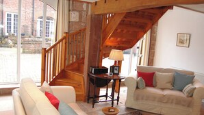 A spacious living room with a wooden staircase, a white sofa, a beige sofa, and a small table with a lamp.