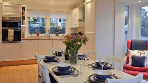 A modern kitchen with white cabinetry, a dining area with blue tableware, and a view of a stone wall through the window. - 3 bedroom accommodation in Rothesay, Isle of Bute (Rothesay, Isle of Bute)
