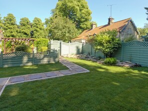 A well-maintained garden with a stone pathway, a wooden pergola, and a stone house with a tiled roof.
