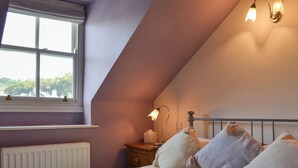 Slaapkamer met schuine plafond, een bed met witte lakens, meerdere kussens, een nachtkastje met een lamp en een raam met uitzicht op bomen.