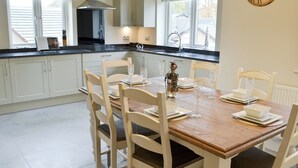 Une cuisine avec des meubles blancs, une table à manger en bois dressée avec de l’assiette blanche, et une horloge au mur.