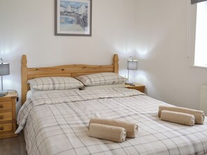 A neatly made bed with a plaid bedspread, two pillows, and rolled towels on a wooden headboard. There is a nightstand with a lamp and a drawer. - A peaceful haven with a summerhouse and just minutes from Mevagissey and Fowey. (St Austell)