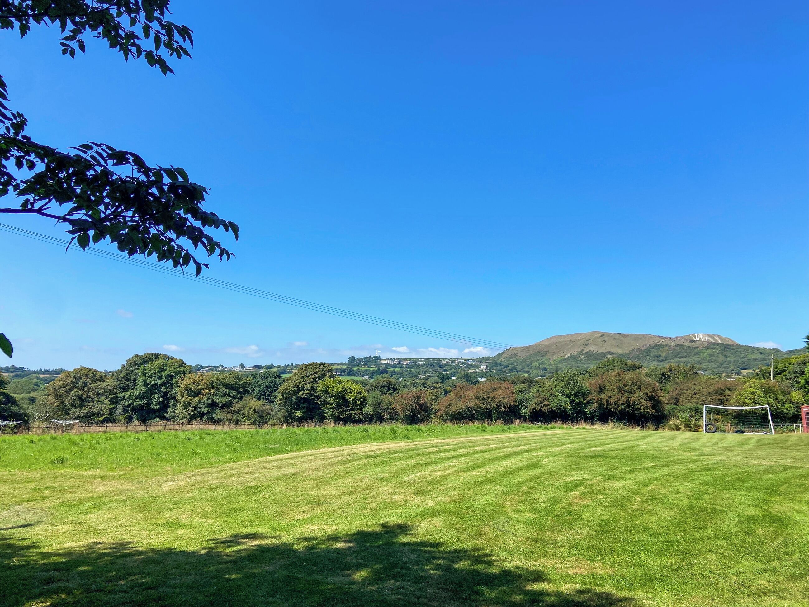 A grassy field with a soccer goal, trees, and a hill in the background.