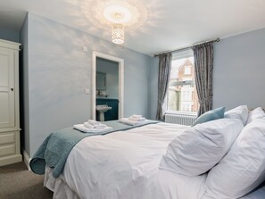 Une chambre à coucher avec un lit, une fenêtre avec des rideaux, un radiateur et une salle de bain visible à travers une porte ouverte.