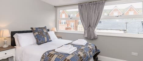 A bedroom with a bed, a nightstand, a lamp, a window with curtains, and a view of buildings outside.
