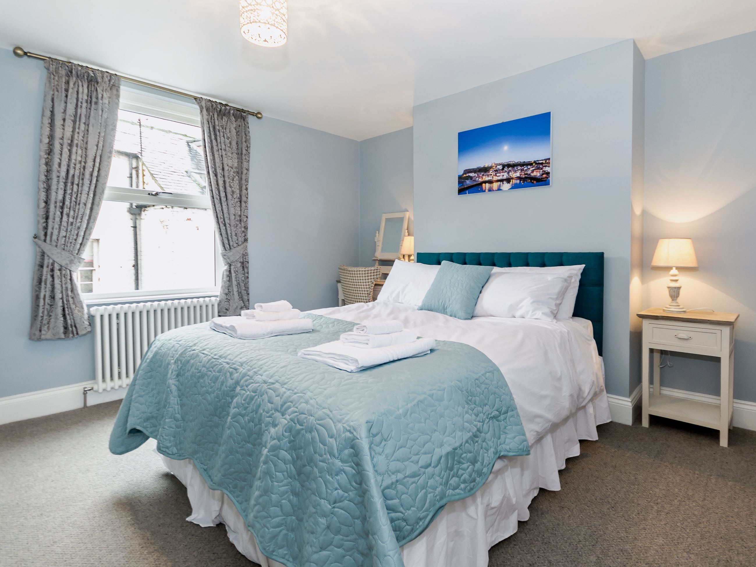 A neatly made bed with a teal quilt, a white pillow, and a teal headboard. There is a window with curtains, a nightstand with a lamp, and a framed picture on the wall.
