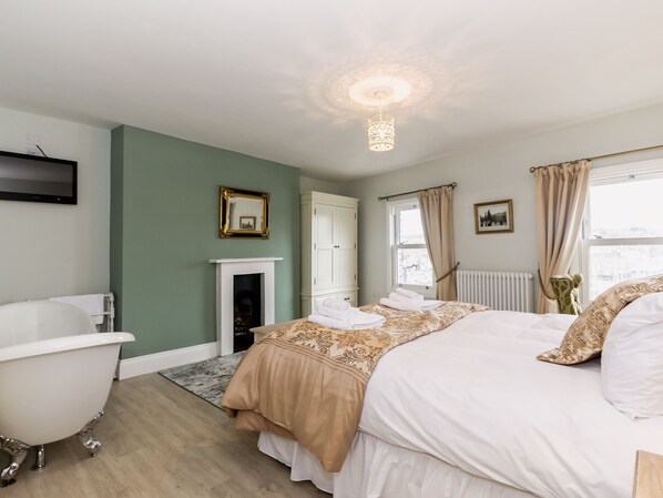 A bedroom with a white bed, a white bathtub, a fireplace, and a window with curtains.