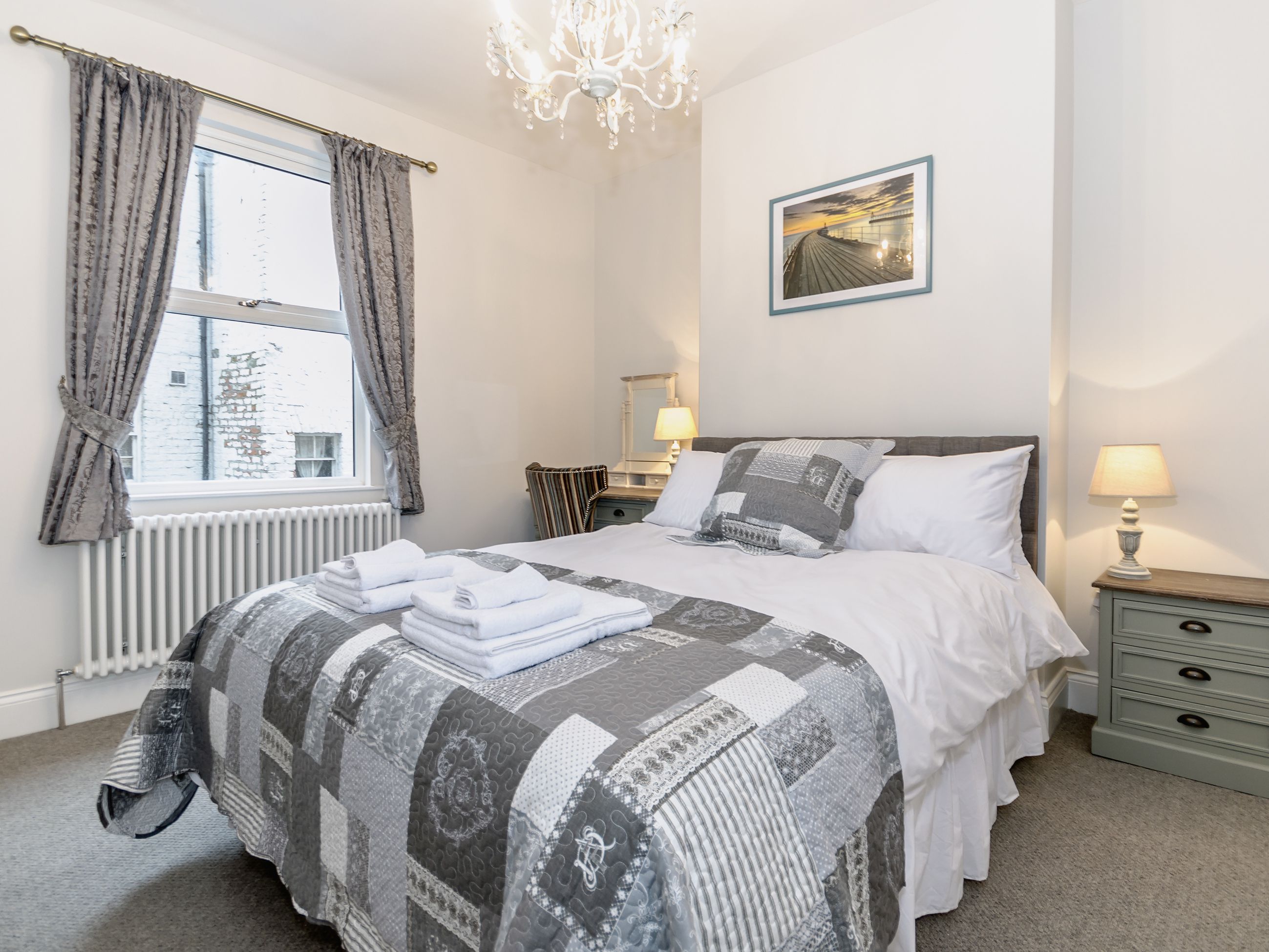 A neatly arranged bedroom with a bed, bedside tables, a radiator, and a window with curtains.