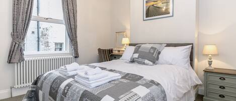 A neatly arranged bedroom with a bed, bedside tables, a radiator, and a window with curtains.