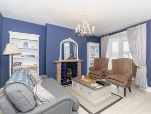 Un salon comprenant un canapé, un fauteuil, une cheminée, une bibliothèque et un lustre.