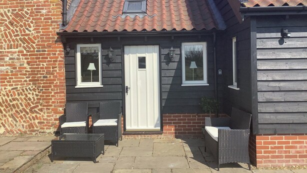 A small house with a tiled roof, a brick wall, and a white door. There are two windows with white frames and a small skylight. Outside, there are two wicker tables with chairs.