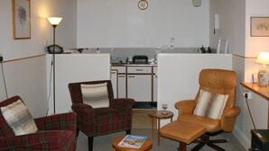 Un espacio habitable pequeño con kitchenette, dos sillones de cuadros, un sillón de cuero y una pequeña mesa con una revista.