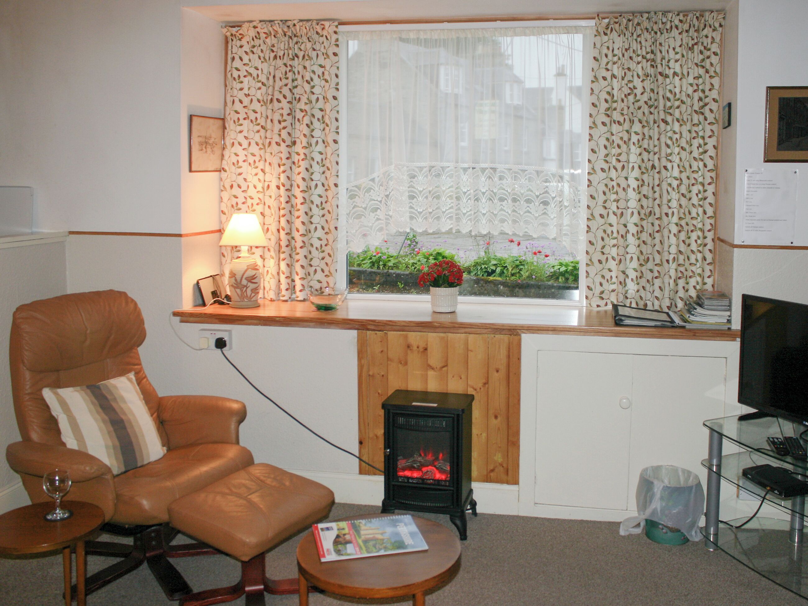Una habitación acogedora con un sillón de cuero, una mesita con un vaso, una chimenea y una ventana con cortinas.