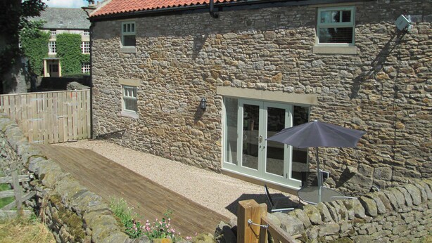 Un bâtiment en pierre avec un toit recouvert de tuiles rouges, une terrasse en bois et un espace patio aménagé avec une table et un parasol.
