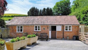 A brick building with a tiled roof, a wooden door, and a small porch area with a table and chairs. - Pig Sty Cottage - E1867 (West Milton)