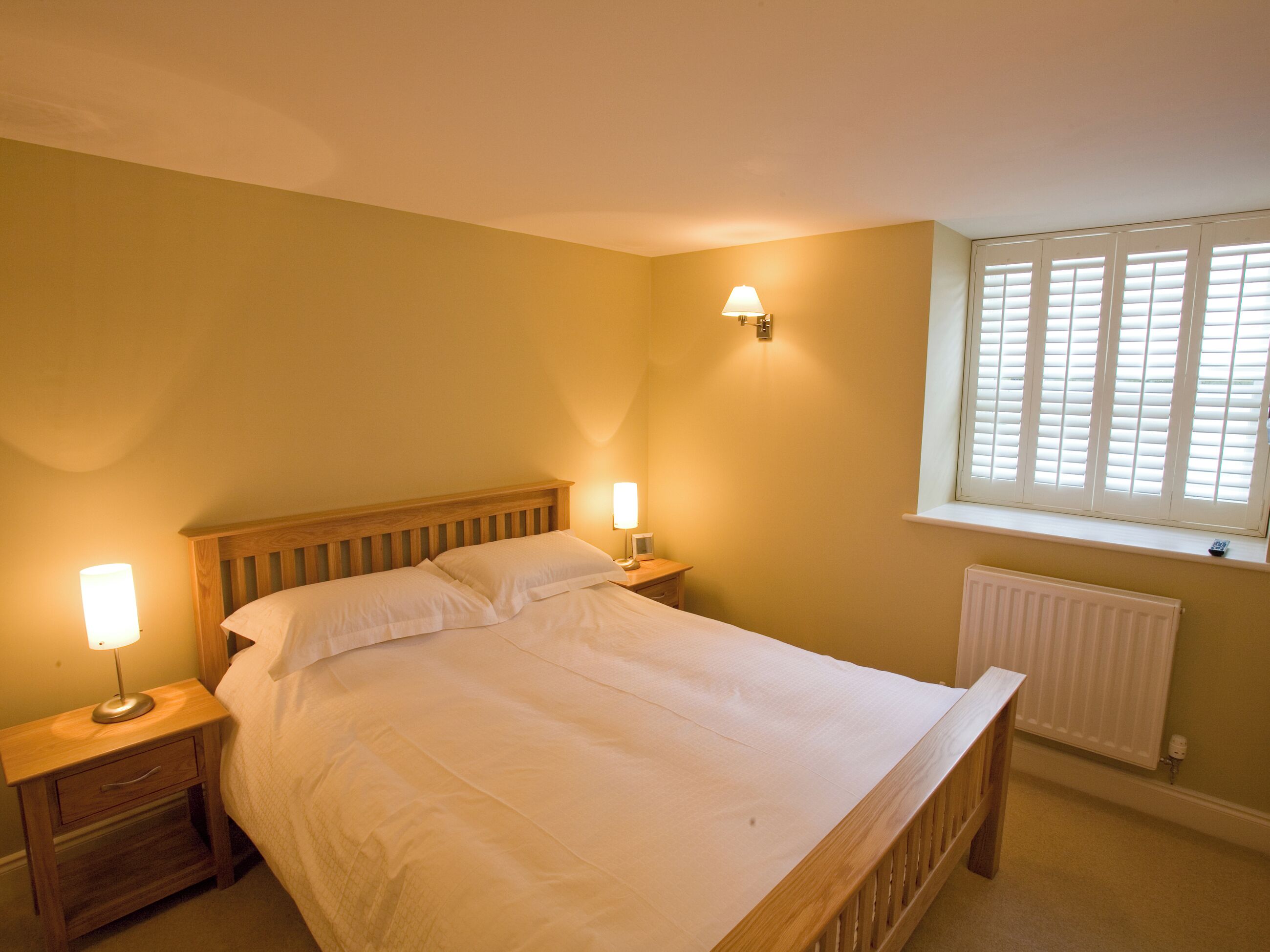 A bedroom with a wooden bed, two bedside lamps, a window with blinds, and a radiator.