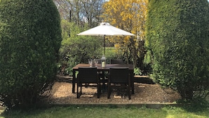 Eine Terrrasse im Freien mit Tisch und Stühlen, umgeben von ordentlich geschnittenen Hecken und einem gelb blühenden Baum.