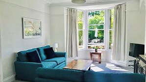 Ein Wohnzimmer mit einem türkisfarbenen Sofa, einem kleinen Tisch, einem Fernseher und einem Fenster mit Vorhängen.