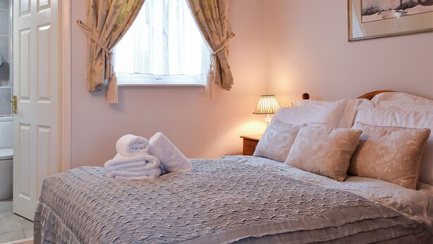 Un dormitorio con cama, almohadas, una lámpara de noche y un ventanal con cortinas.
