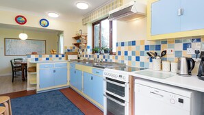 Een keuken met blauwe kastjes, een fornuis, een oven en een vaatwasser. Er is een raam met uitzicht op bomen.