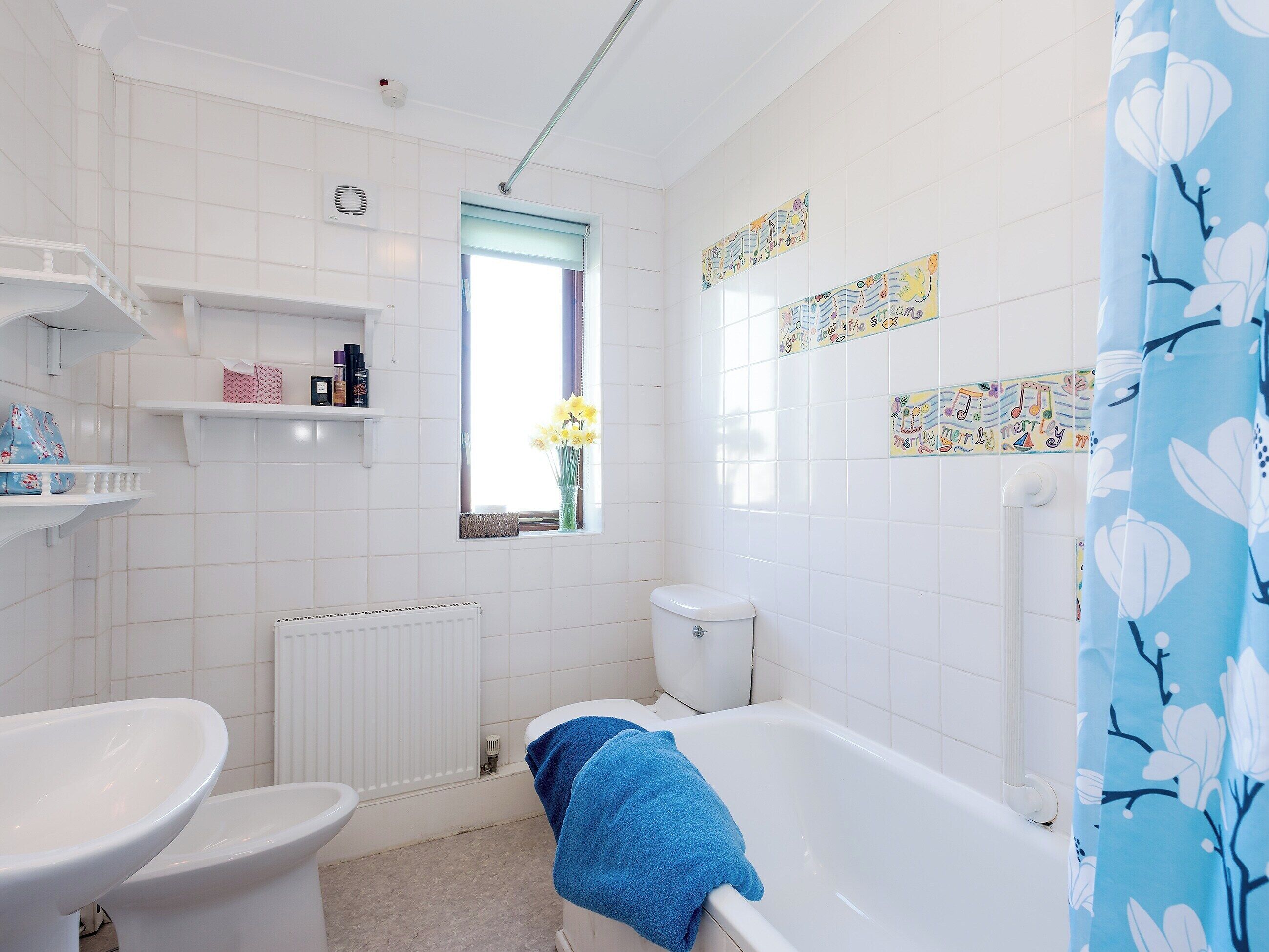 A bathroom with a toilet, bidet, and bathtub. There is a window with a view of flowers outside, and the walls are decorated with colorful tiles.