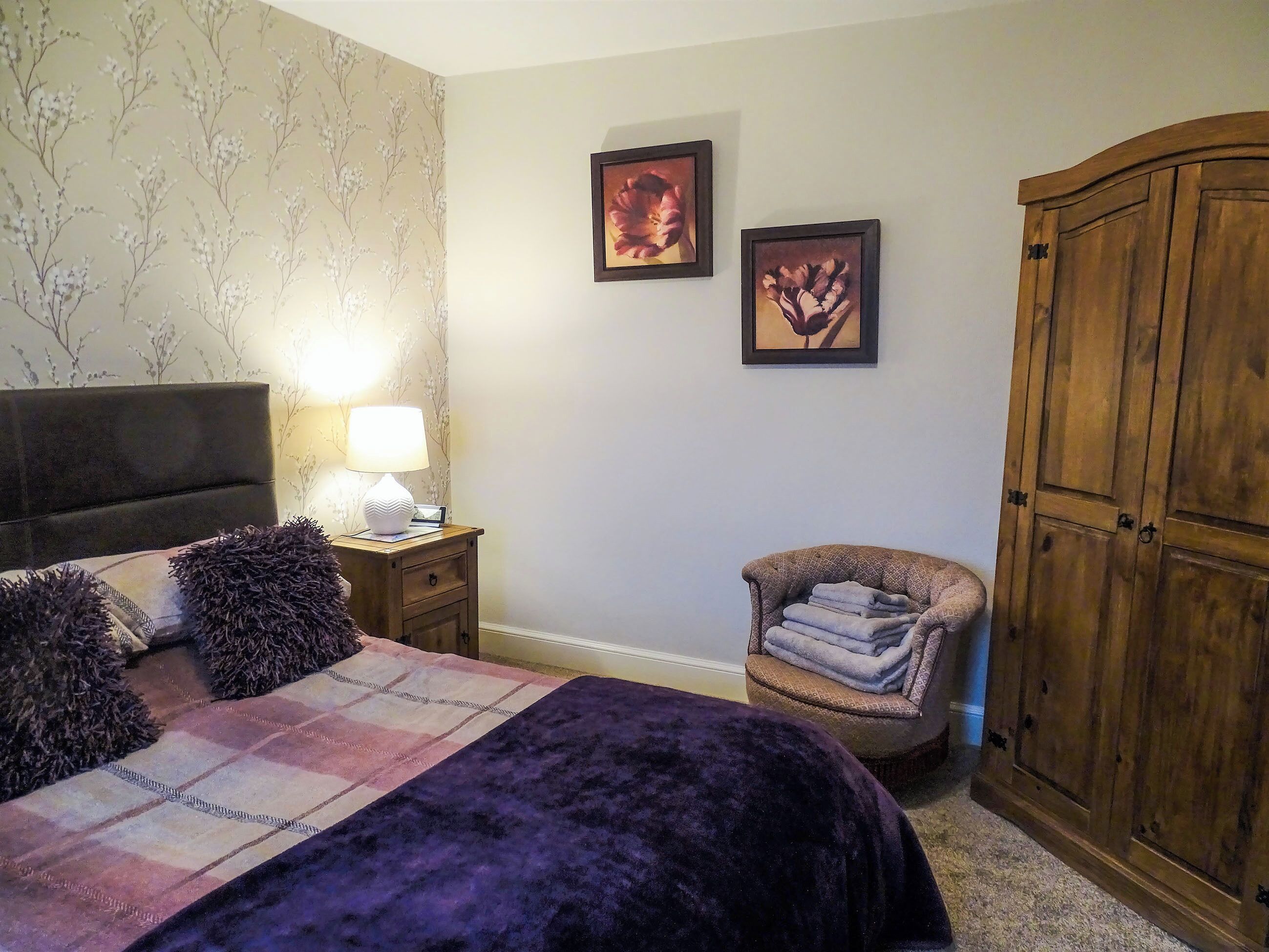 A bedroom with a bed, a nightstand with a lamp, a chair with a blanket, and two framed pictures on the wall.