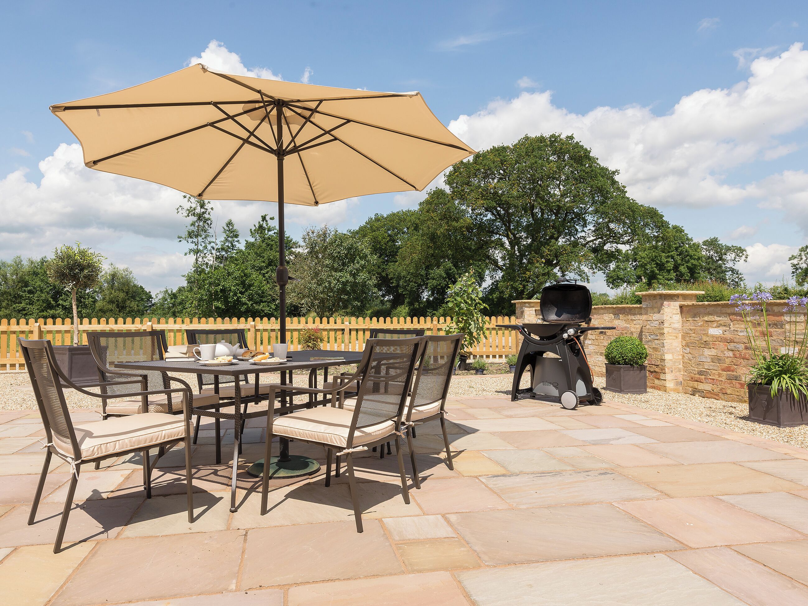 Een terras buiten met een tafel, stoelen, een grote parasol en een barbecue.