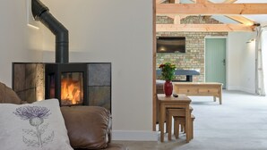 Een woonkamer met een open haard, een bank, een klein tafeltje met een vaas, en uitzicht op een keuken met een bakstenen muur en houten balken.